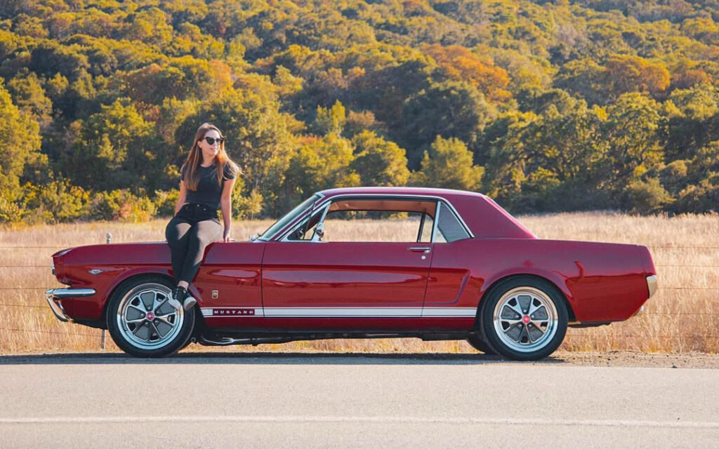 Francesca sitting on the front left fender of her 1966 Ford Mustang on a California country road.