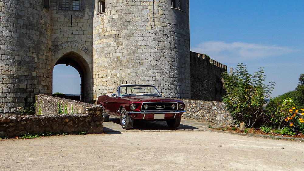 1967 Mustang Convertible Vintage Burgundy parked in front of a medieval castle in France, blending American muscle with European heritage